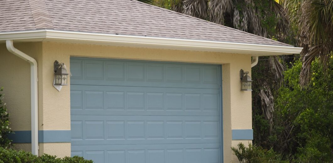 A closed light blue garage door on a beige house with two outdoor wall lights and greenery surrounding the driveway.