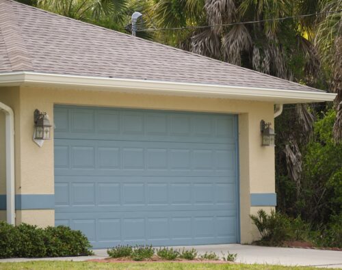 A closed light blue garage door on a beige house with two outdoor wall lights and greenery surrounding the driveway.