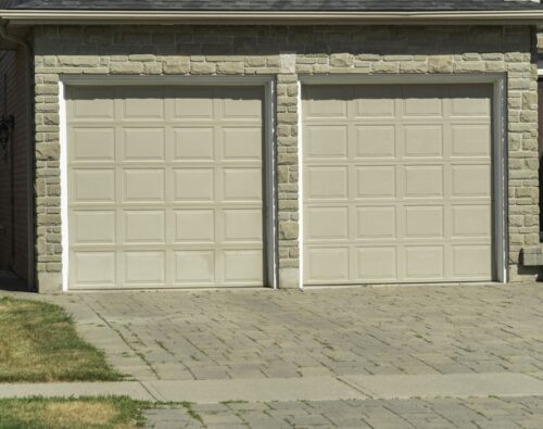 Two closed beige garage doors on a stone-faced building with a paved driveway and some patches of grass in the foreground.