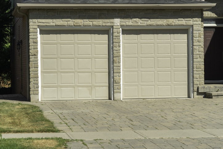 Two closed beige garage doors on a stone-faced building with a paved driveway and some patches of grass in the foreground.