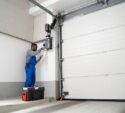 A technician in blue overalls repairs or installs a garage door opener using tools, with a toolbox set on the ground nearby.