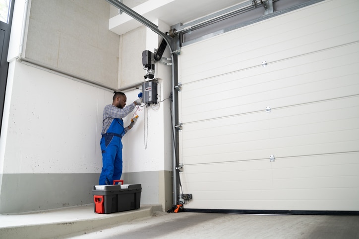 A technician in blue overalls repairs or installs a garage door opener using tools, with a toolbox set on the ground nearby.