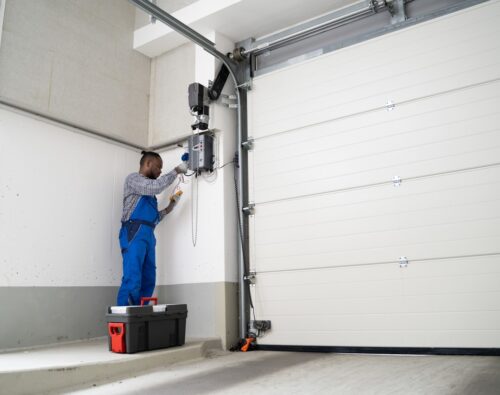 A technician in blue overalls repairs or maintains an electric garage door opener, standing next to a large closed garage door with a toolbox on the ground.