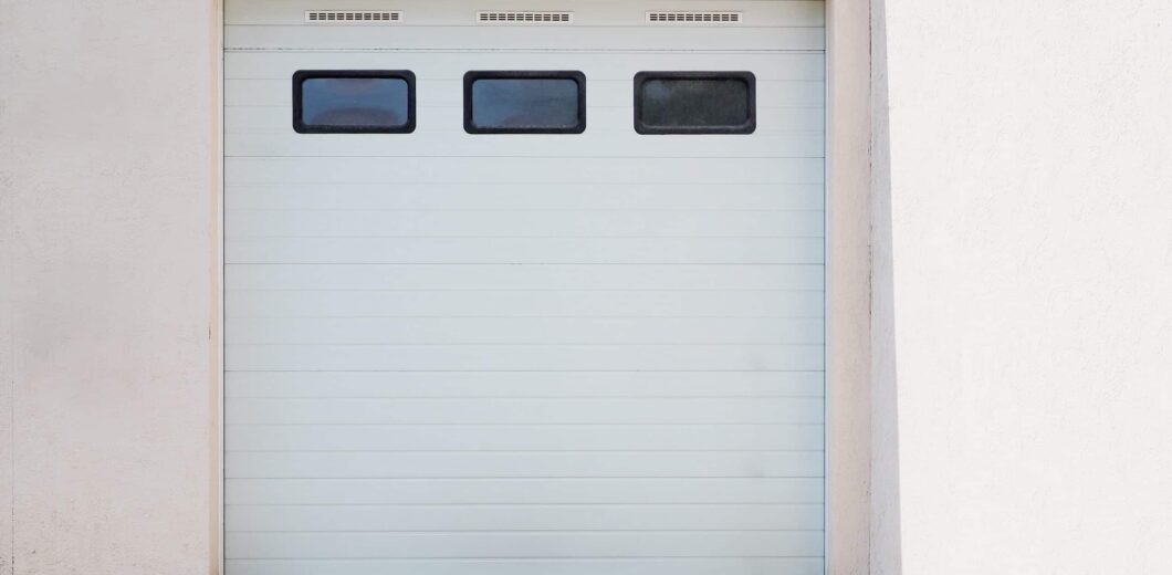 Closed white garage door with three rectangular windows near the top, set in a light-colored exterior wall on a concrete driveway.