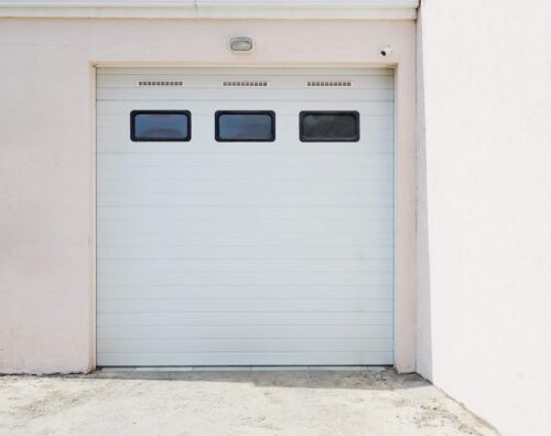 Closed white garage door with three rectangular windows near the top, set in a light-colored exterior wall on a concrete driveway.
