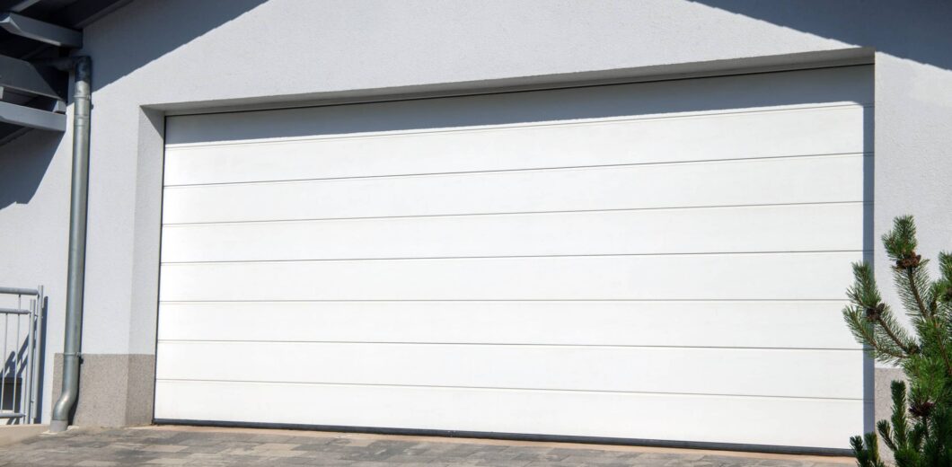 A closed white sectional garage door on a modern house with a paved driveway and a small evergreen shrub in the foreground.