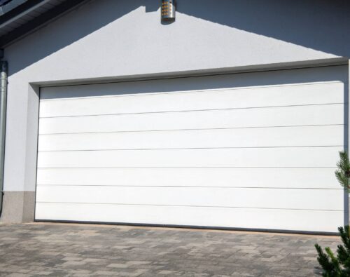 A closed white sectional garage door on a modern house with a paved driveway and a small evergreen shrub in the foreground.