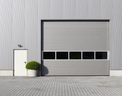 Gray industrial building exterior with a large closed garage door, a small side door, a wall-mounted light, and a potted shrub on a paved surface.