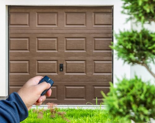 A person holds a remote control in front of a closed brown garage door, with greenery visible in the foreground.