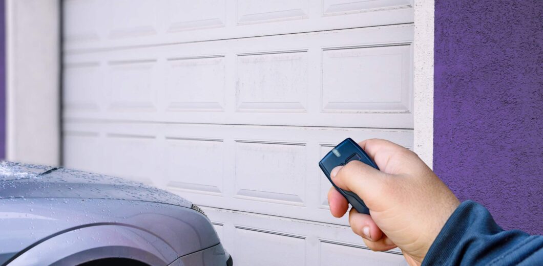 A person uses a remote control to open a closed white garage door next to a parked silver car.