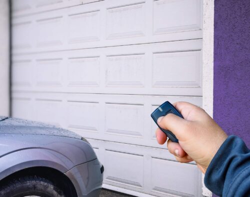 A person uses a remote control to open a closed white garage door next to a parked silver car.