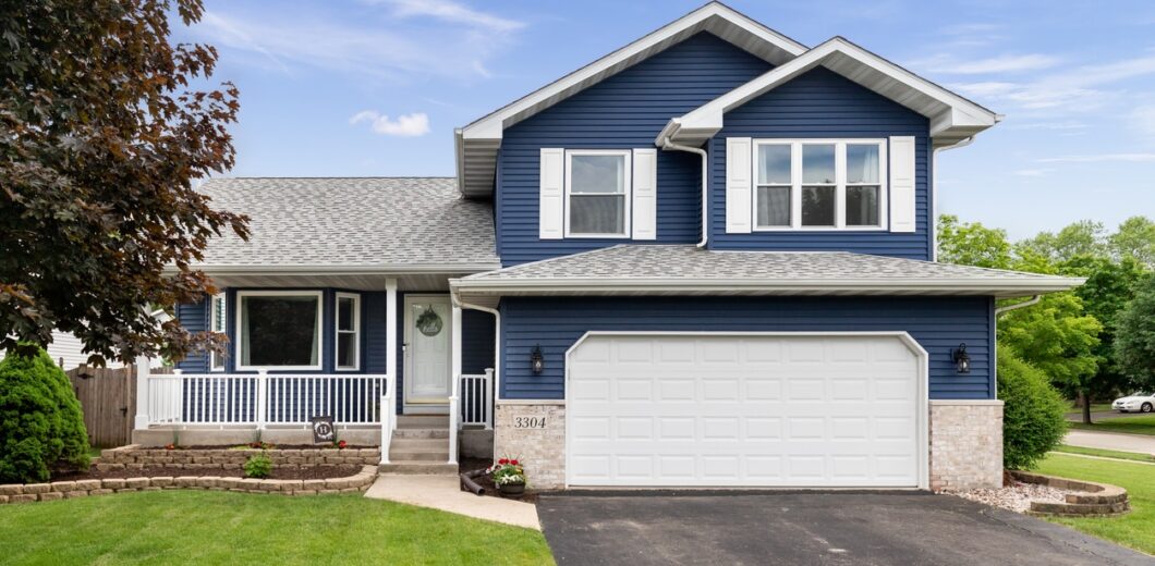 Two-story blue house with white trim, a front porch, attached two-car garage, paved driveway, green lawn, and trees in the yard, under a partly cloudy sky.