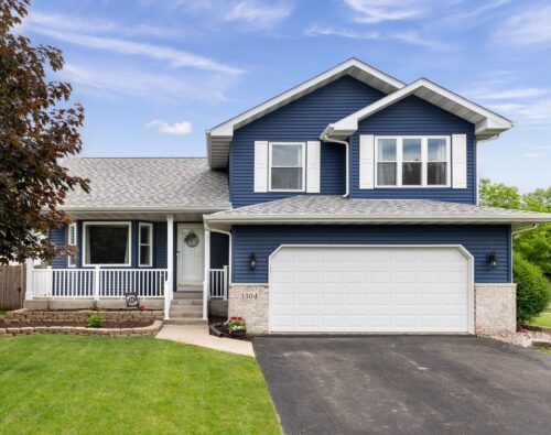 Two-story blue house with white trim, a front porch, attached two-car garage, paved driveway, green lawn, and trees in the yard, under a partly cloudy sky.