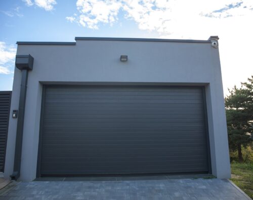 A modern gray garage with a closed overhead door, an outdoor light above, and trees visible to the right under a partly cloudy sky.