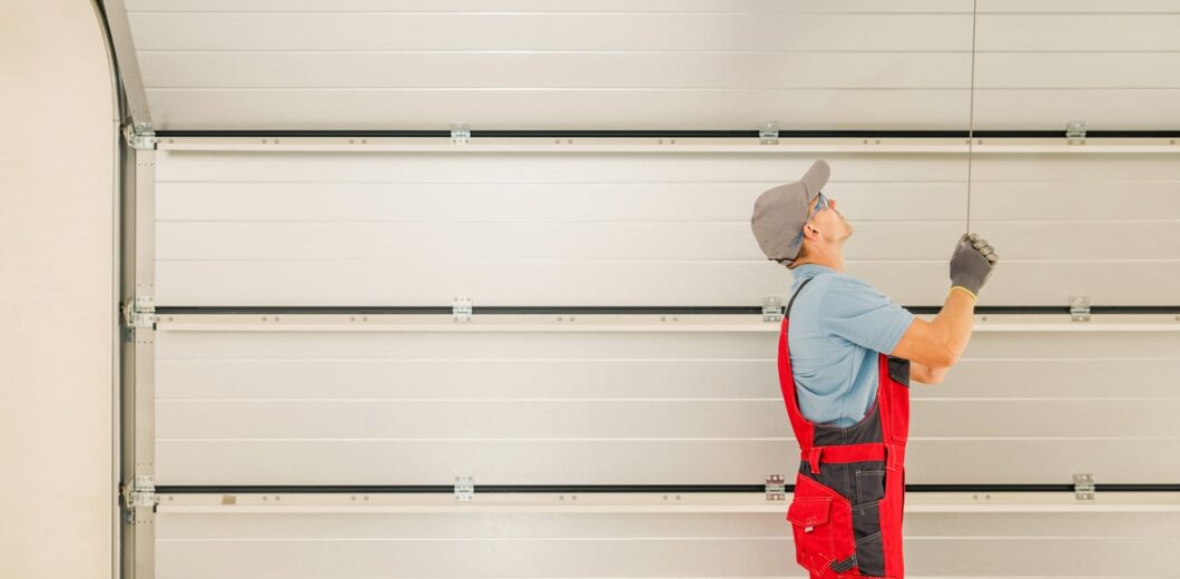 A worker in red overalls and a cap is pulling a cord to open or close a large white garage door.