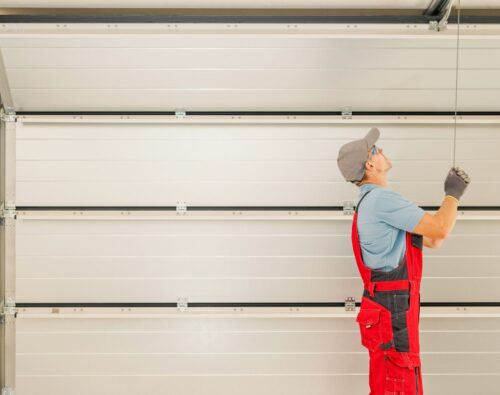 A worker in red overalls and a cap is pulling a cord to open or close a large white garage door.