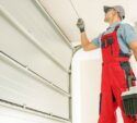 A technician in red overalls and gloves repairs a garage door while holding a toolbox.