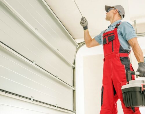 A technician in red overalls and gloves repairs a garage door while holding a toolbox.