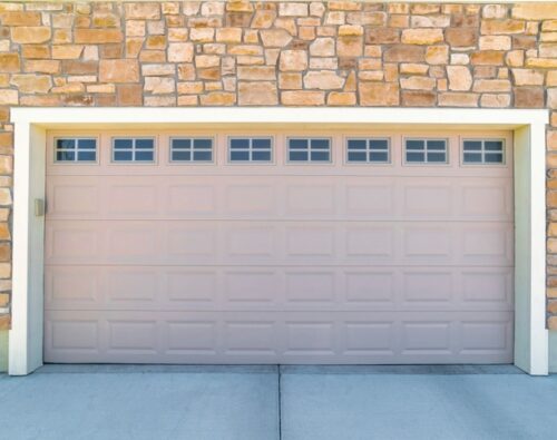 Closed beige garage door set in a stone wall facade with small windows at the top and two black lantern-style wall lights on either side.