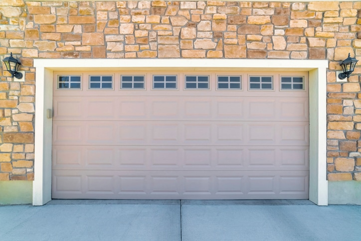 Closed beige garage door set in a stone wall facade with small windows at the top and two black lantern-style wall lights on either side.