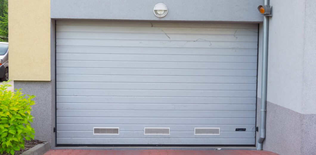 Closed gray garage door set in a building with a yellow and gray facade, brick pavement, and a small green shrub to the left.