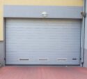 Closed gray garage door set in a building with a yellow and gray facade, brick pavement, and a small green shrub to the left.