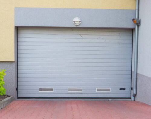 Closed gray garage door set in a building with a yellow and gray facade, brick pavement, and a small green shrub to the left.