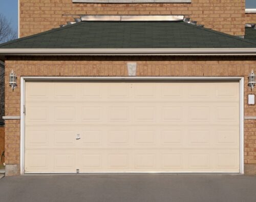 A closed beige double garage door attached to a brick house, with two lantern-style lights on either side and a concrete driveway in front.