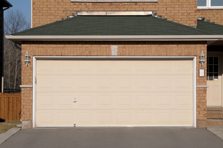 A closed beige double garage door attached to a brick house, with two lantern-style lights on either side and a concrete driveway in front.