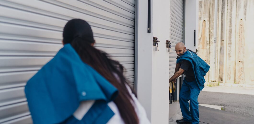 Two workers in blue uniforms are opening or closing metal roll-up doors on a loading dock or storage facility.