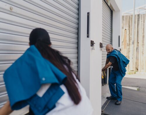 Two workers in blue uniforms are opening or closing metal roll-up doors on a loading dock or storage facility.