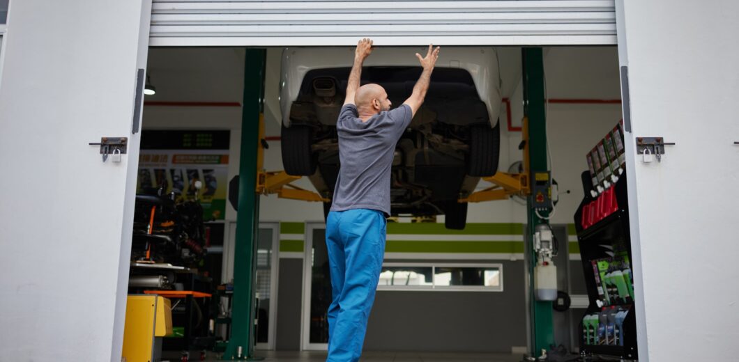 A person in blue pants raises a garage door at an auto shop, with a car lifted on a hydraulic platform inside.