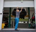 A person in blue pants raises a garage door at an auto shop, with a car lifted on a hydraulic platform inside.