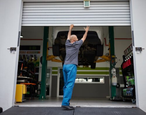 A person in blue pants raises a garage door at an auto shop, with a car lifted on a hydraulic platform inside.