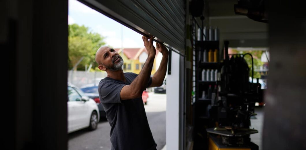 A man stands inside a garage, manually lifting a metal roll-up door. Cars and trees are visible outside in the background.