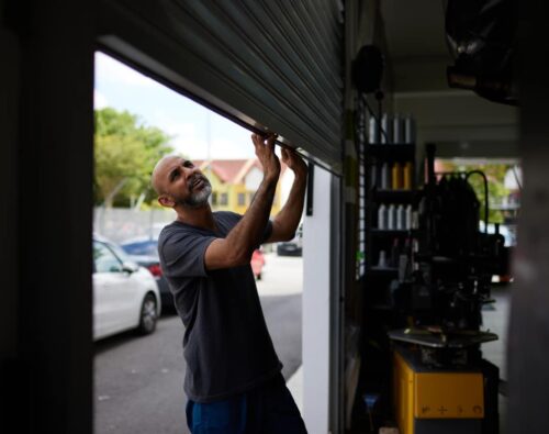 A man stands inside a garage, manually lifting a metal roll-up door. Cars and trees are visible outside in the background.