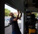 A man lifts a metal garage door at an auto repair shop, with cars and buildings visible outside in the background.