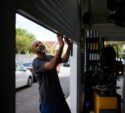 A man lifts a metal garage door inside an automotive workshop, with parked cars and buildings visible outside in daylight.