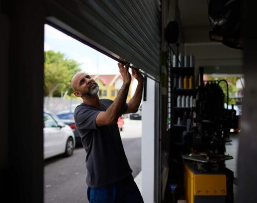 A man lifts a metal garage door at an auto repair shop, with cars and buildings visible outside in the background.