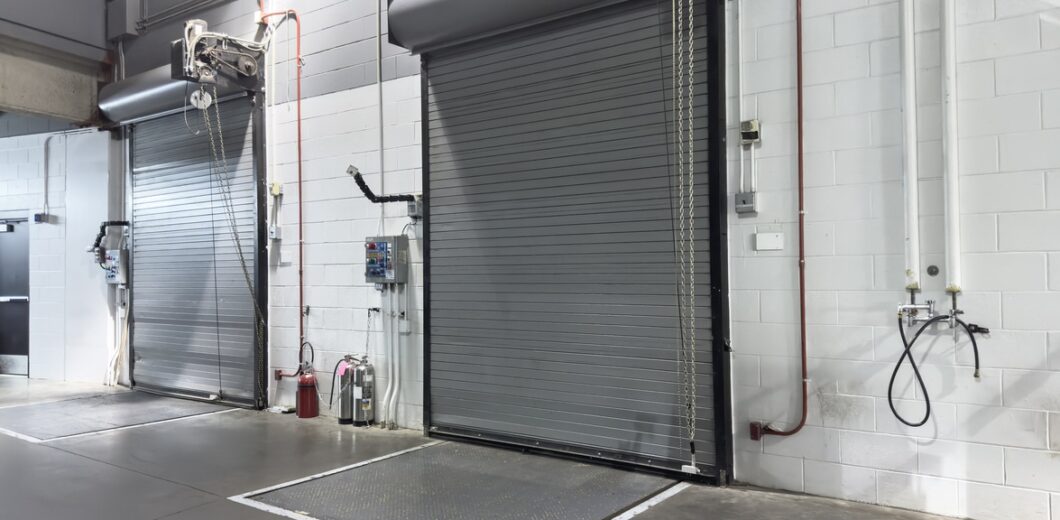 Two gray industrial roll-up doors in a warehouse setting, with wall-mounted control panels, a fire extinguisher, and utility fixtures nearby.