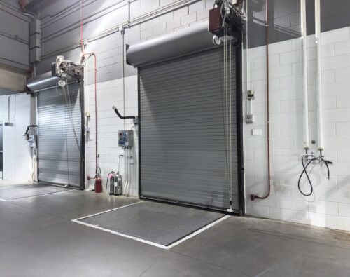 Two gray industrial roll-up doors in a warehouse setting, with wall-mounted control panels, a fire extinguisher, and utility fixtures nearby.