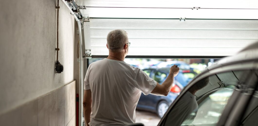 A man with gray hair in a white t-shirt stands in a garage, using a remote to open the garage door with parked cars visible outside.