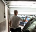 A man with gray hair in a white t-shirt stands in a garage, using a remote to open the garage door with parked cars visible outside.