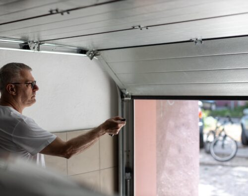 A man in a white t-shirt uses a remote control to open a garage door, with a bicycle and car visible outside.
