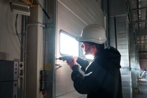 A worker wearing a hard hat uses a power drill to install or repair a metal door inside an industrial building.