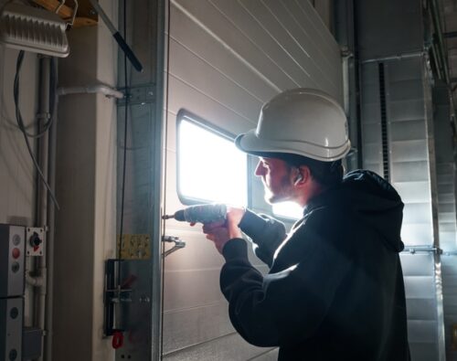 A worker wearing a hard hat uses a power drill to install or repair a metal door inside an industrial building.