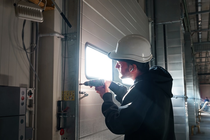 A worker wearing a hard hat uses a power drill to install or repair a metal door inside an industrial building.