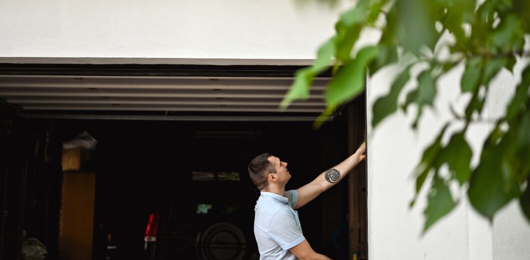 A man in a light blue shirt stands outside a garage, reaching up to close or open the garage door. A black vehicle is parked inside. Green leaves are visible in the foreground.