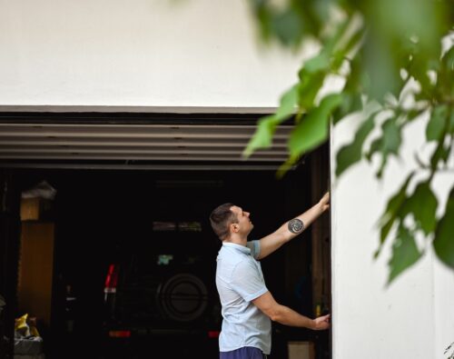 A man in a light blue shirt stands outside a garage, reaching up to close or open the garage door. A black vehicle is parked inside. Green leaves are visible in the foreground.
