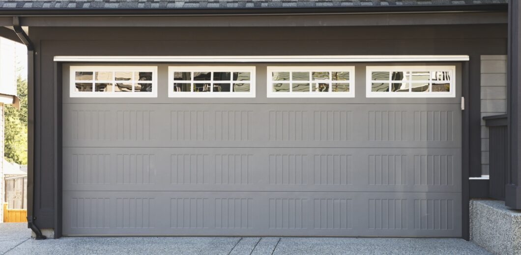 Gray residential garage door with rectangular panel design and a row of small square windows at the top, set in a modern house exterior.